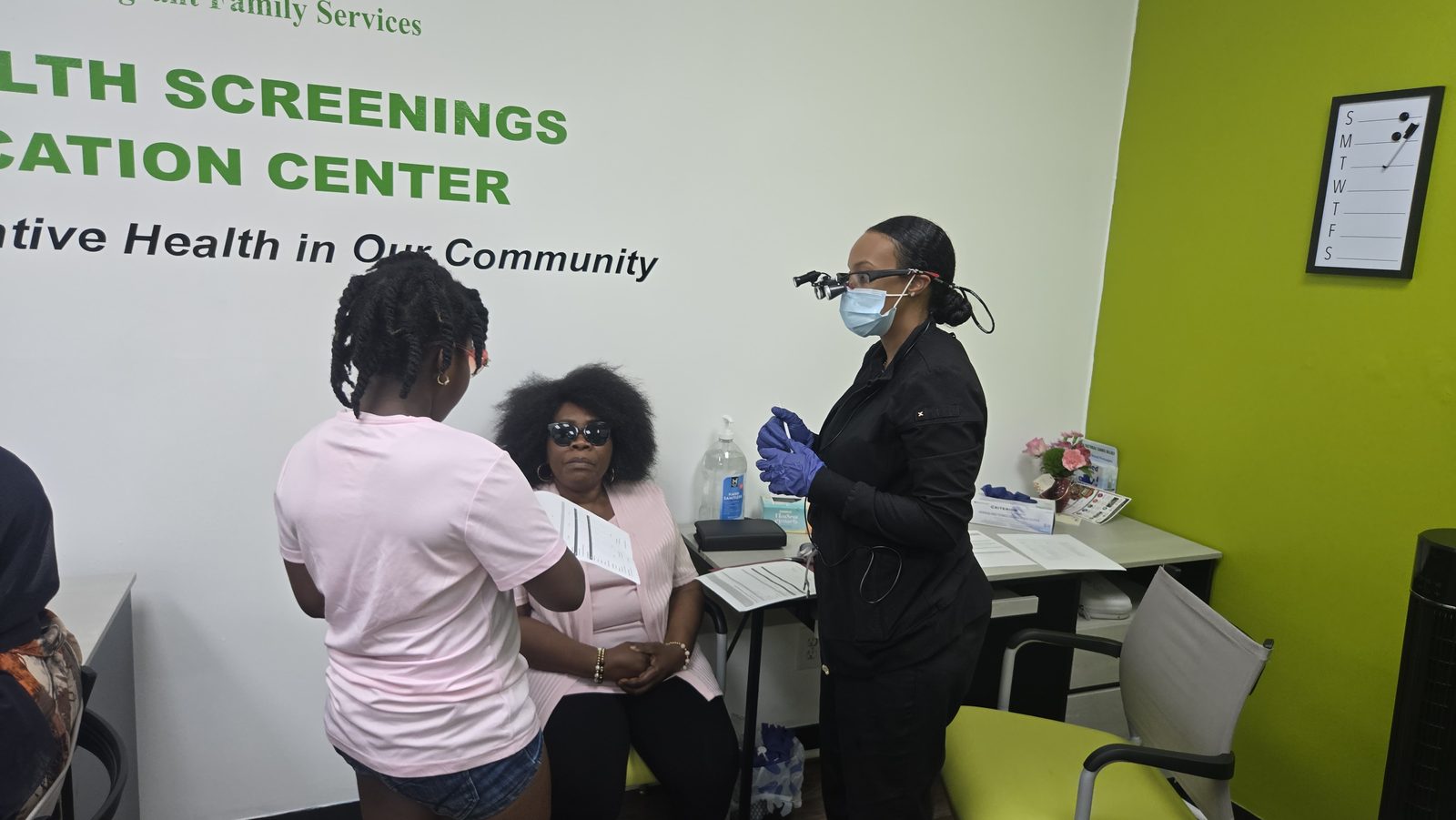 A healthcare worker speaks with seated adults in a room with AIFS signage.